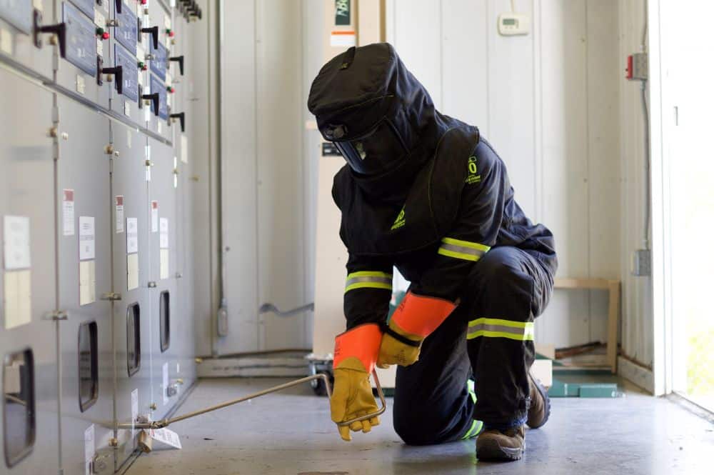 A man in a safety suit kneels to inspect a panel, showcasing qualified electrical worker training in action.