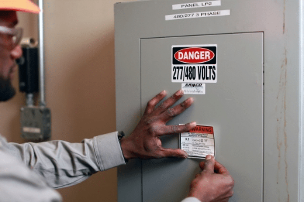 A man in an orange safety vest conducts an arc flash hazard analysis while labeling a box.